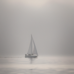 Sailboat cuts through fog-shrouded waters at dawn, its white sail billowing forward while heavy mist obscures the distant horizon